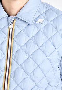 Veste matelassée bleu clair avec col, fermeture éclair à rayures multicolores et épingle logo blanche sur la poitrine.