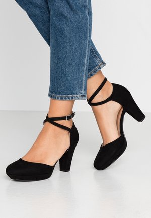 Zwarte suède hoge hakken met een ronde neus, enkelbandjes en een chunky hak, gecombineerd met opgerolde blauwe denim jeans.