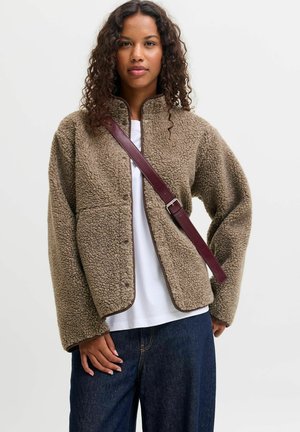 Femme aux cheveux bouclés porte une veste sherpa marron sur une chemise blanche, un jean bleu foncé et une bandoulière en cuir marron.