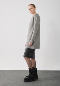 Frau, die im Profil steht und einen grauen, übergroßen Pullover, einen schwarzen, knielangen Lederrock, transparente Strumpfhosen und schwarze Schnürstiefel trägt.