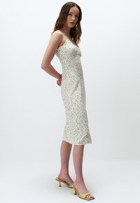Blumiges Midikleid in Weiß mit gelben und blauen Blumen, ausgestattet mit Spaghettiträgern und einer figurbetonten Silhouette. Tragen mit gelben Absatzsandalen.