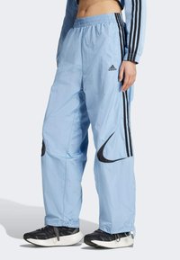 Ljusblå joggingbyxor med svarta Nike- och Adidas-logotyper, elastisk midja, sidfickor och svarta ränder längs sidorna.