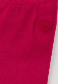 Helles pinkes Gewebe mit subtiler Textur und einem kleinen, runden Logo mit einem stilisierten Design in der unteren linken Ecke.