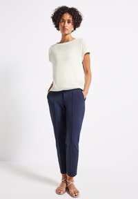 Světle krémový pletený top s krátkým rukávem v kombinaci s přizpůsobenými tmavomodrými kalhotami, které mají boční kapsy a slim fit střih. Vzhled doplňují metalické sandály na pásky.