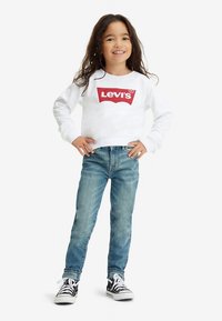 Λευκό φούτερ με κόκκινο λογότυπο Levi's, σε συνδυασμό με ανοιχτόχρωμο μπλε τζιν και μαύρα-λευκά αθλητικά παπούτσια. Κανονική εφαρμογή, μακριά μανίκια.