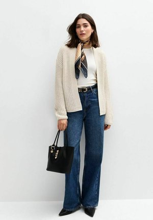 Un cardigan en maille crème sur un haut blanc, associé à un jean bleu évasé. Tenant un sac fourre-tout noir avec des accessoires dorés et une écharpe accentuée.