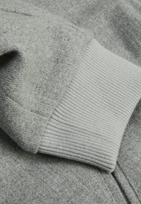 Sivi duksericasti materijal s rebrastim manžetama; meka tekstura i ravnomjeran pleteno uzorak, s blagim heather izgledom i podesivim zatvaranjem na patentni zatvarač.