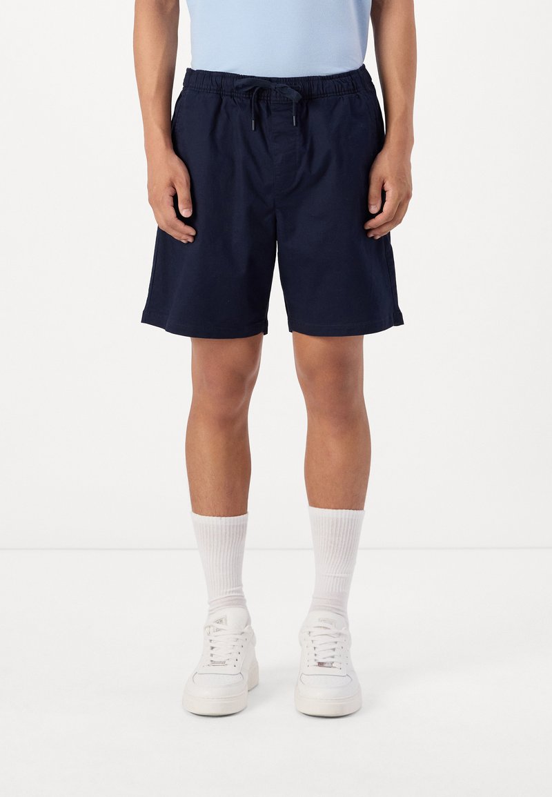 Shorts de bain bleu marine avec taille élastique et cordon de serrage. Associés à des chaussettes blanches et des baskets. Tissu lisse et coupe au genou.