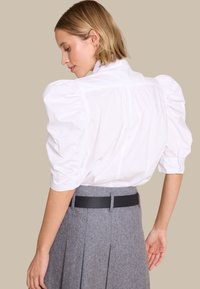 Hvit bluse med puffede ermer og høy krage, med knappeclosure, slitt med et grått plisserte skjørt og et sort texturert belte.