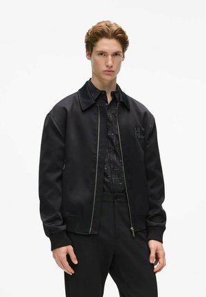 Joven con el pelo corto castaño claro, vestido con una camisa negra abotonada, chaqueta negra con cremallera y pantalones negros, de pie frente a un fondo blanco.