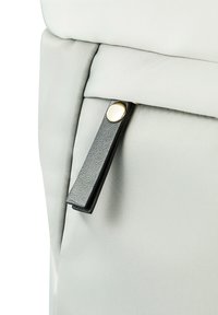 Détail d'un sac à dos en tissu gris présentant un tirant de fermeture éclair noir texturé et un bouton-pression doré. Surface lisse avec de subtils motifs de couture.