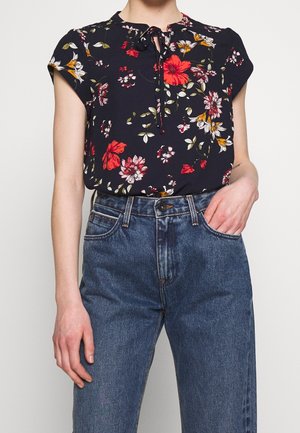 Persona que lleva una blusa negra con flores rojas y blancas, metida dentro de unos vaqueros azules de talle alto, con una mano en el bolsillo.