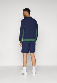 Uomo che indossa una maglietta a maniche lunghe blu navy e verde, pantaloni corti blu navy, calze bianche e scarpe da ginnastica bianche, in piedi di fronte a un muro.