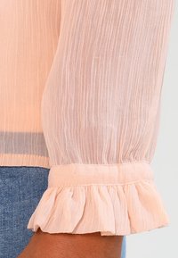 Blouse légère rose pâle en tissu transparent texturé et plissé avec des poignets à volants. La manche présente un design doux et fluide, ajoutant un détail délicat.