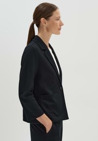 Zwarte blazer met een gesneden kraag, twee voorzakken en een enkele knoopsluiting. Lichtgewicht stof met een subtiele textuur.