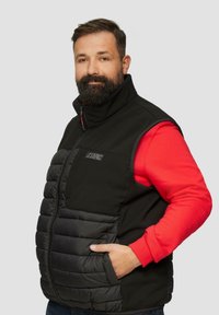 Schwarze isolierte Weste mit einem Puffdesign, hohem Kragen und Fronttasche. Getragen über einem roten Langarmshirt mit Logo-Details.