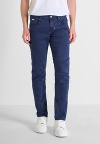 Jeans ajustados de color azul marino con una textura suave, que presentan un diseño de cinco bolsillos y un cierre de botón, combinados con zapatillas blancas.