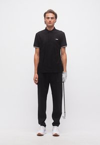 Νεαρός άνδρας που φοράει μαύρο πόλο και παντελόνι, λευκά αθλητικά παπούτσια και γάντι γκολφ, κρατώντας ένα γκολφ ρόπαλο σε απλό λευκό φόντο.