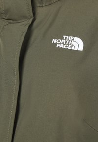Olive zielony materiał o gładkiej fakturze, zdobiony białym haftowanym logo "The North Face" w lewym górnym rogu.