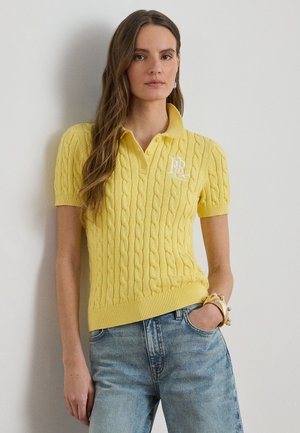 Ženska v rumeni polo majici s kabelski vzorec in belim logotipom ter svetlomodrih kavbojkah stoji ob enostavni svetli steni.