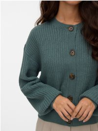 Roheline kootud kardigan ümara kaelusega, millel on suured pruunid nööbid ja sooniline muster. Lahked varrukad ulatuvad randmeteni.