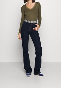 Olivfarbenes, geripptes Langarmshirt mit Spitzen-Ausschnitt, kombiniert mit dunkelblauen Schlagjeans und gemustertem Gürtel. Blaue und weiße Sneakers vervollständigen das Outfit.