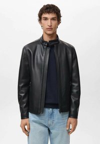 Giacca di pelle nera con colletto alto, zip frontale e texture sottile. Indossata sopra un maglione scuro e jeans azzurri chiari.