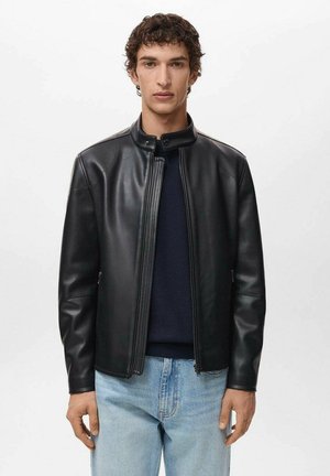 Giacca di pelle nera con colletto alto, zip frontale e texture sottile. Indossata sopra un maglione scuro e jeans azzurri chiari.