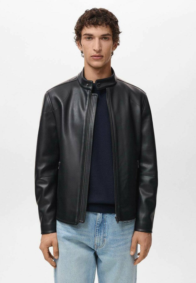Giacca di pelle nera con colletto alto, zip frontale e texture sottile. Indossata sopra un maglione scuro e jeans azzurri chiari.