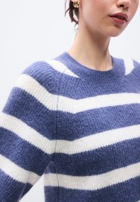 Pulover cu dungi albastre și albe, cu decolteu rotund, confecționat din material tricotat moale. Are un croi lejer și țesătură texturată.