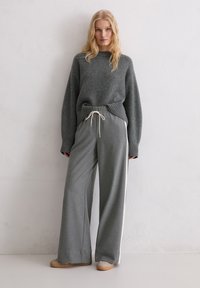 Blondynka ubrana w szary, oversize'owy sweter oraz szerokie szare spodnie z białymi paskami po bokach, stojąca przy białej ścianie.