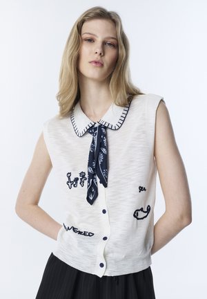 Jonge vrouw met blond haar, gekleed in een mouwloos wit gilet met blauwe borduursels en een blauw sjaaltje met patroon dat onder de kraag gestrikt is.