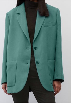 Persoon draagt een losse blauwgroene blazer met twee knopen over een zwarte coltrui en donkere broek, staand tegen een effen lichte achtergrond.