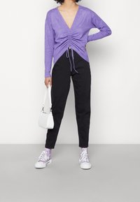 Fioletowa bluzka z długim rękawem z głębokim dekoltem w serek i marszczeniem w talii, zestawiona z czarnymi spodniami, białymi trampkami i małą białą torebką.