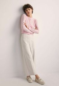 Jasnoróżowy krótki kardigan z zapięciem na guziki, zestawiony z białymi spodniami w stylu szerokich nogawek i beżowymi loafersami z akcentem w postaci łańcuszka.