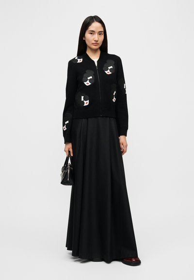 Crni kardigan na patentni zatvarač s umjetničkim aplikacijama lica, uparen s lepršavom crnom maxi suknjom i malom crnom torbicom. Glatka tekstura tkanine.