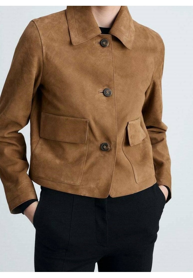 Veste en daim marron boutonnée avec large col et poches à rabat sur le devant, portée sur un pantalon noir, mains dans les poches.