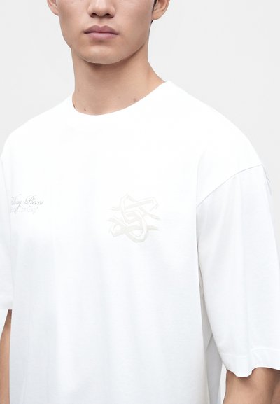 Νέος άνδρας που φοράει λευκό φαρδύ T-shirt με διακριτικά κεντημένα λογότυπα στο στήθος, σε ουδέτερο φόντο.