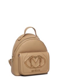 Mochila de cuero marrón claro con forma redondeada, que cuenta con un bolsillo delantero con un acento de logo metálico y un asa superior para llevarla. Cierre con cremallera.