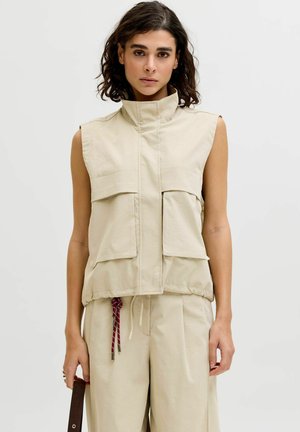 Žena s tamnom kosom do ramena nosi bež prsluk bez rukava s prednjim džepovima i odgovarajuće bež hlače, držeći tamnoljubičastu traku.