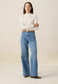 Weißes Spitzenshirt mit langen Ärmeln und hohem Kragen, kombiniert mit hellblauen, hochtaillierten Schlagjeans und einem beigen Gürtel. Texturiertes Material und auffällige Taschen.