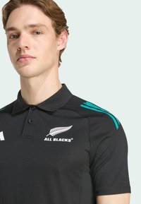 Polo noir orné d'accents turquoise et d'un col structuré. Affiche le logo "All Blacks" et un motif de fougère sur la poitrine.