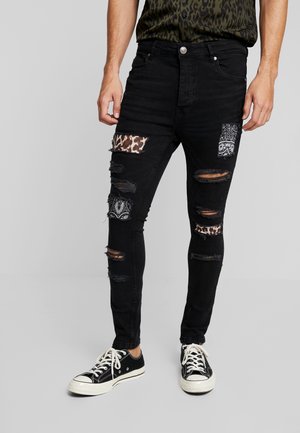 Hombre que lleva vaqueros pitillo negros con parches rotos que muestran inserciones de tela de leopardo y cachemira, combinados con zapatillas negras y blancas.