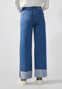 Jeans largos azuis feitos de denim, com uma bainha desfiada e um destaque azul claro. Dois bolsos traseiros são visíveis.