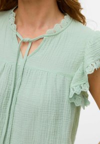 Blusa verde chiaro con un tessuto texturizzato, bordatura in pizzo a scallop al colletto e alle maniche, con un'apertura a chiave legata al collo.