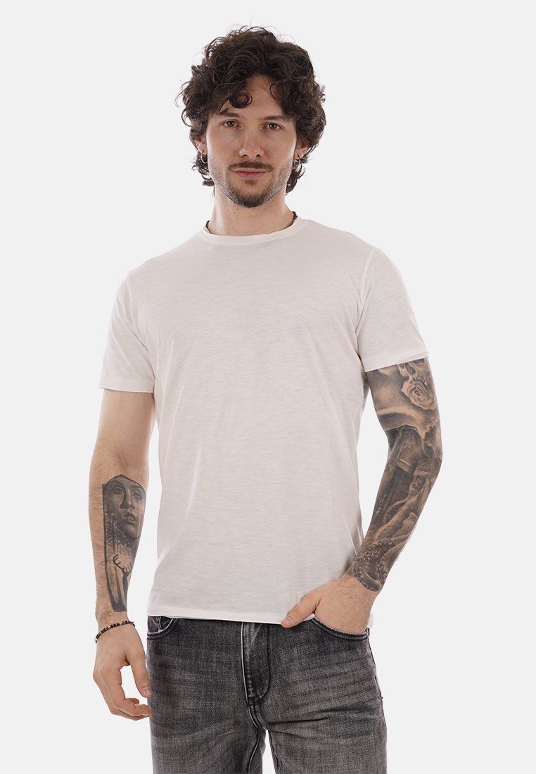 Homme aux cheveux bouclés, barbe, et tatouages sur les bras, portant un t-shirt blanc uni et un jean foncé, debout avec une main dans la poche.