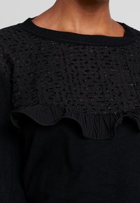 Pull noir avec un empiècement texturé et à motifs, agrémenté de détails volantés. Le matériau est doux et le design comprend un col rond.
