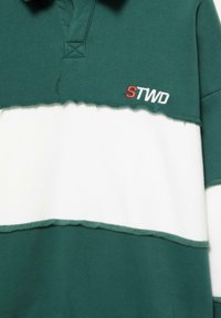 Polo verde e bianco con un design a spacco, caratterizzato da orli grezzi, colletto e logo ricamato "STWD" in rosso sul petto.