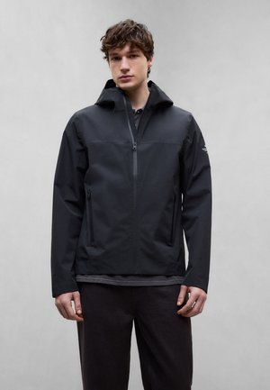 Ung mann iført en svart hettejakke med glidelåslommer, stående mot en enkel lys grå bakgrunn.