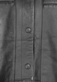 Sort læderstof med en glat tekstur, syede sømme og to runde trykknapper foran, der fremhæver holdbarhed og design detaljer.